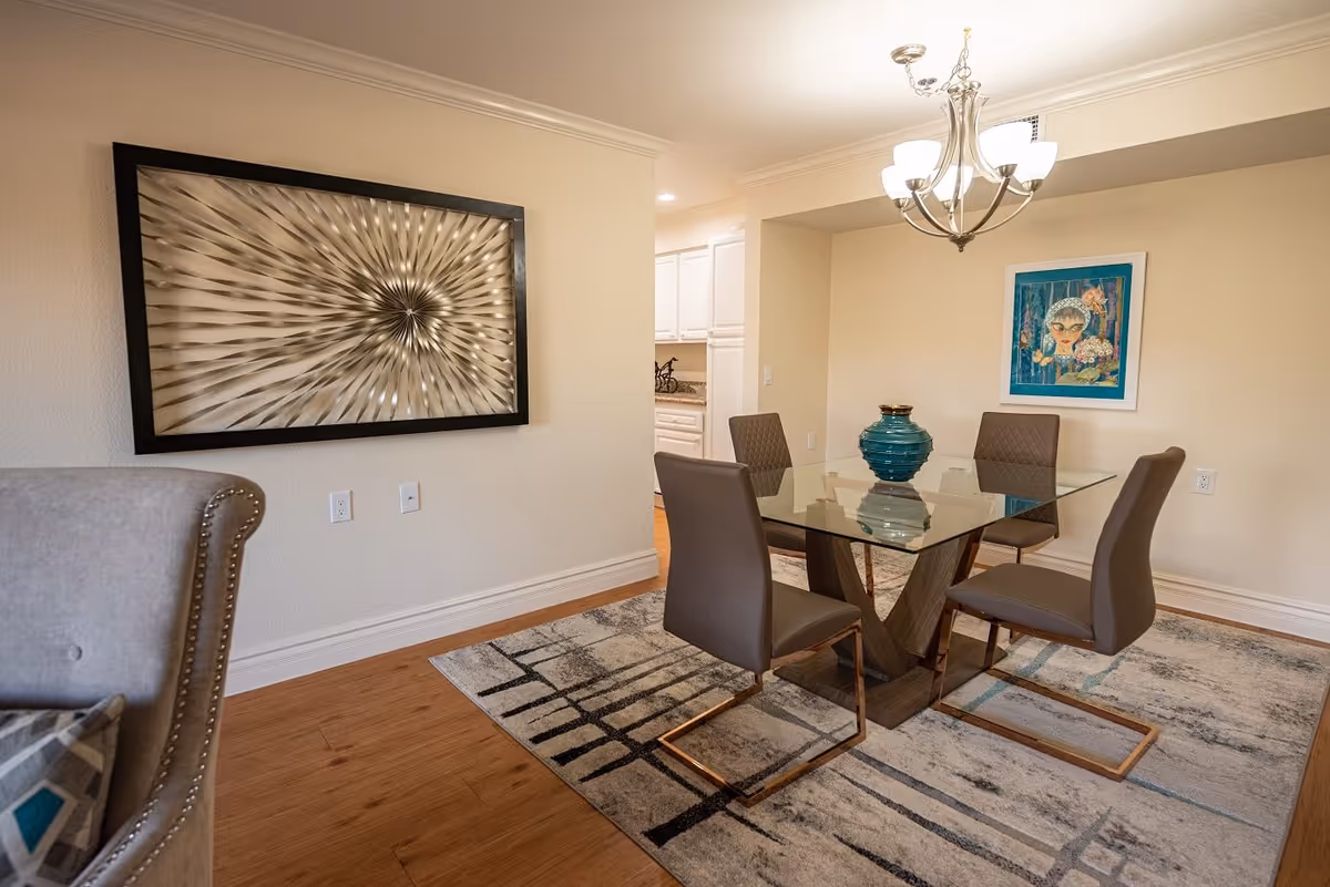 Modern dining room with a glass-top table, four chairs, chandelier, wall art and a decorative vase.