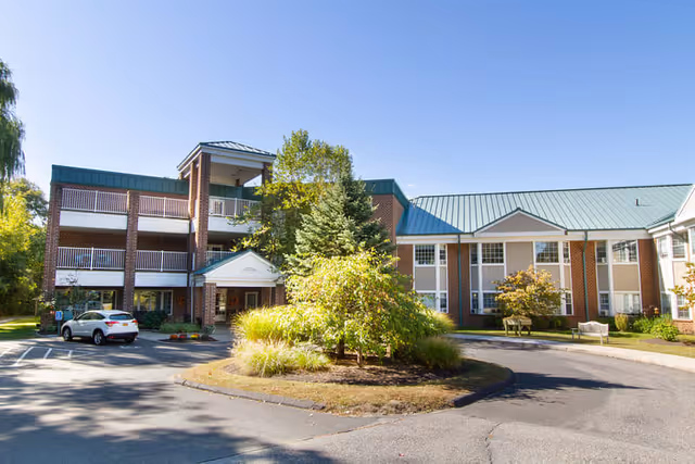 Exterior view of The Cascades Assisted Living facility showing a multi-story brick building with a green metal roof, surrounded by trees and landscaping. There is a circular driveway with a small island of shrubs and grass in the center, and a white car parked near the entrance.