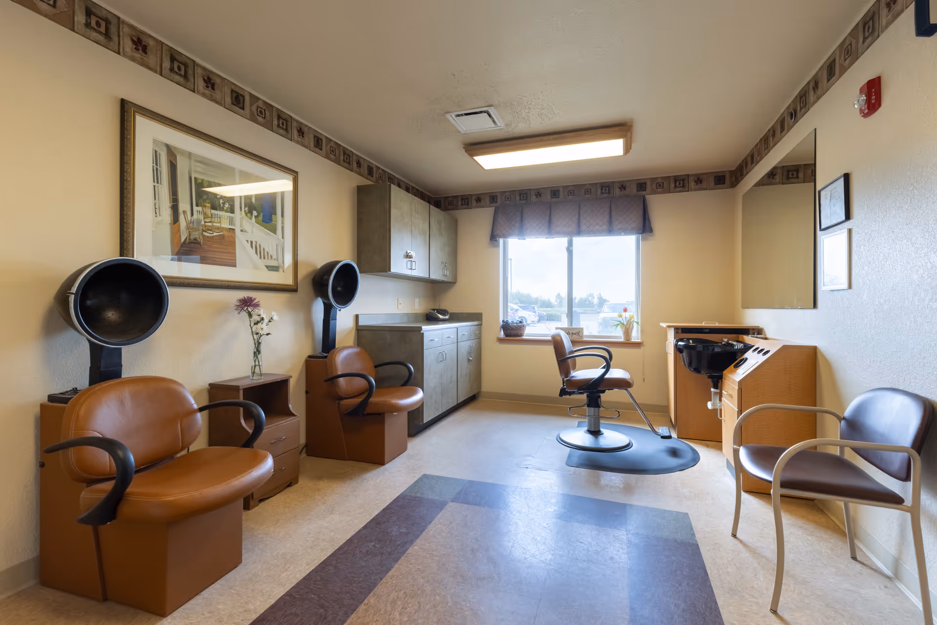 A hair salon room in a senior living facility with styling chairs, hooded dryers, a wash station, cabinets and a window.