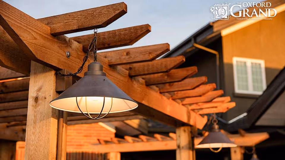 Close-up view of a wooden pergola with hanging industrial-style light fixtures, with a residential building in the background during sunset.