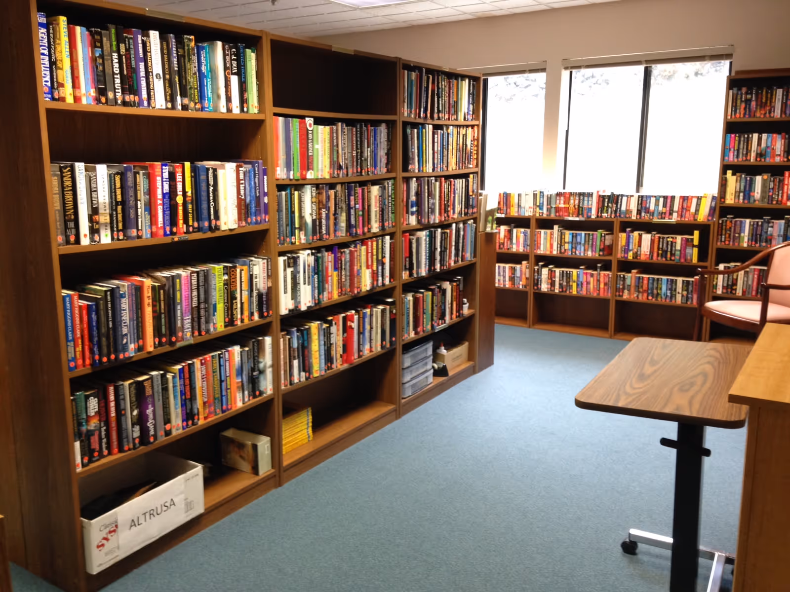 Carpeted reading room with wooden bookshelves filled with books, a table and chair near a window.