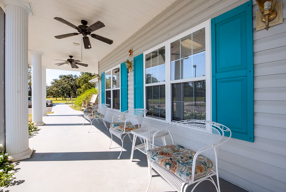 A covered outdoor porch area with white metal chairs and a small table, featuring floral cushions. The porch has large white columns, ceiling fans, and turquoise window shutters on the building wall. Trees and a parking area are visible in the background.