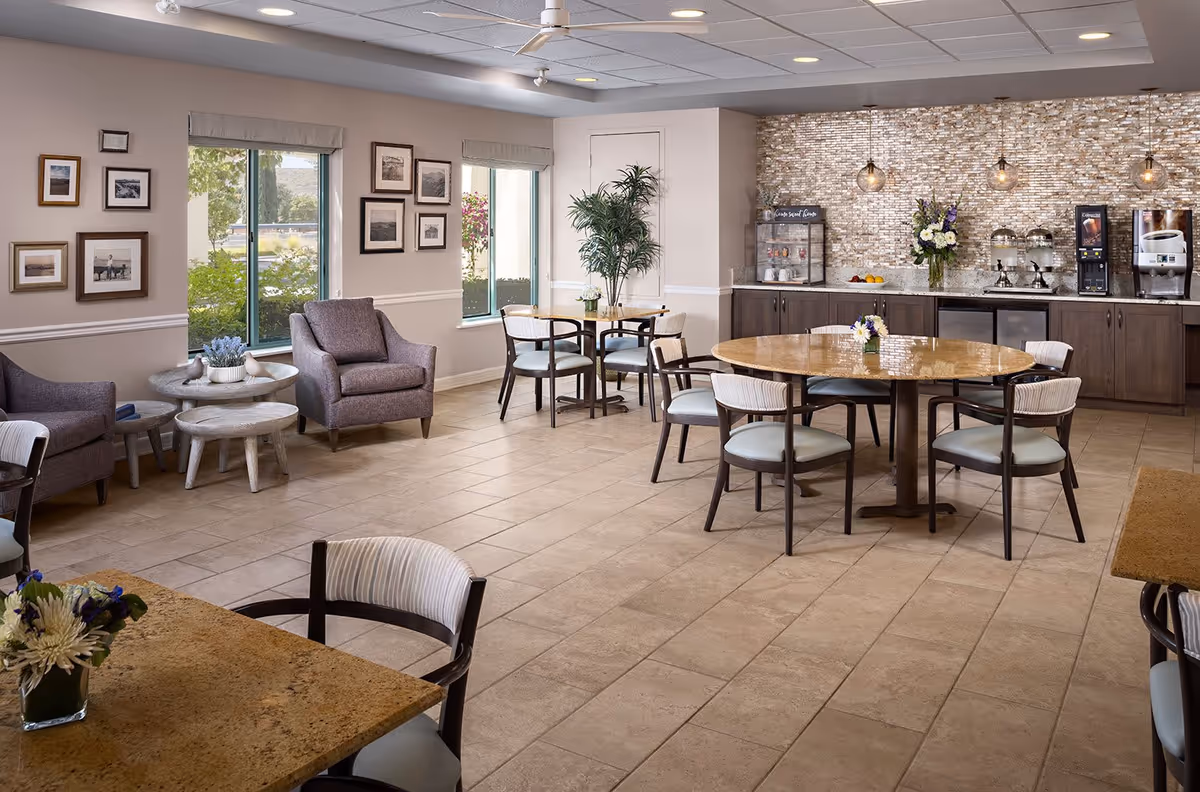 A bright and spacious common area with round tables and chairs arranged for dining or socializing. There are two comfortable armchairs with side tables near windows that let in natural light. The back wall features a countertop with beverage dispensers, a flower arrangement, and a textured stone backsplash. The room has tiled floors and a ceiling fan.