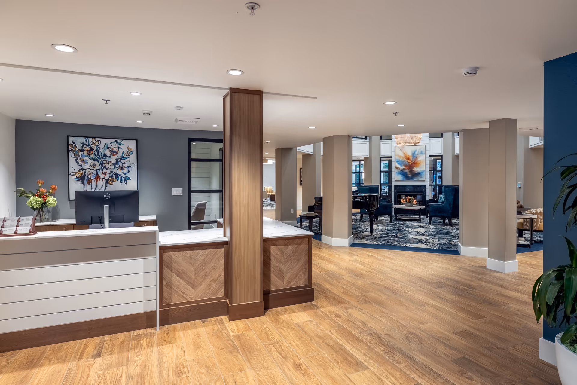Reception area of a senior living facility with a modern front desk, wooden flooring, and a seating area in the background featuring a grand piano, armchairs, and a fireplace with artwork above it.