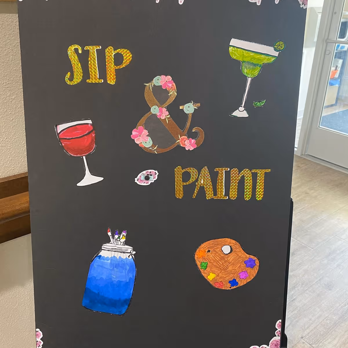 A black poster board with colorful cut-out letters and drawings. The text reads 'SIP & PAINT' with the ampersand decorated with flowers. There are drawings of a red wine glass, a green margarita glass with a lime wedge, a blue jar holding paintbrushes, and a painter's palette with colorful paint blobs. The poster is displayed indoors near a door with a window.