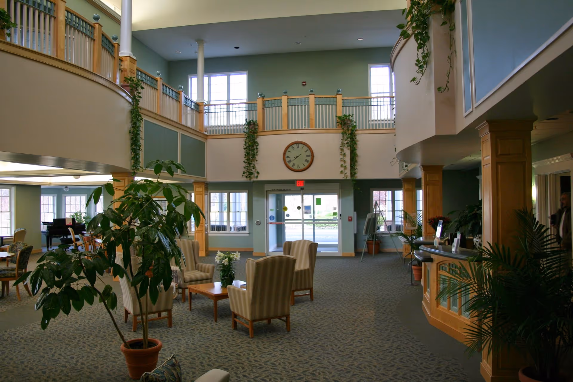 Spacious senior living facility lobby with high ceilings and large windows. The area features comfortable armchairs arranged around a coffee table, potted plants, and a reception desk on the right. A large clock is mounted on the wall above the entrance doors, and there is a balcony railing on the upper level with hanging greenery.