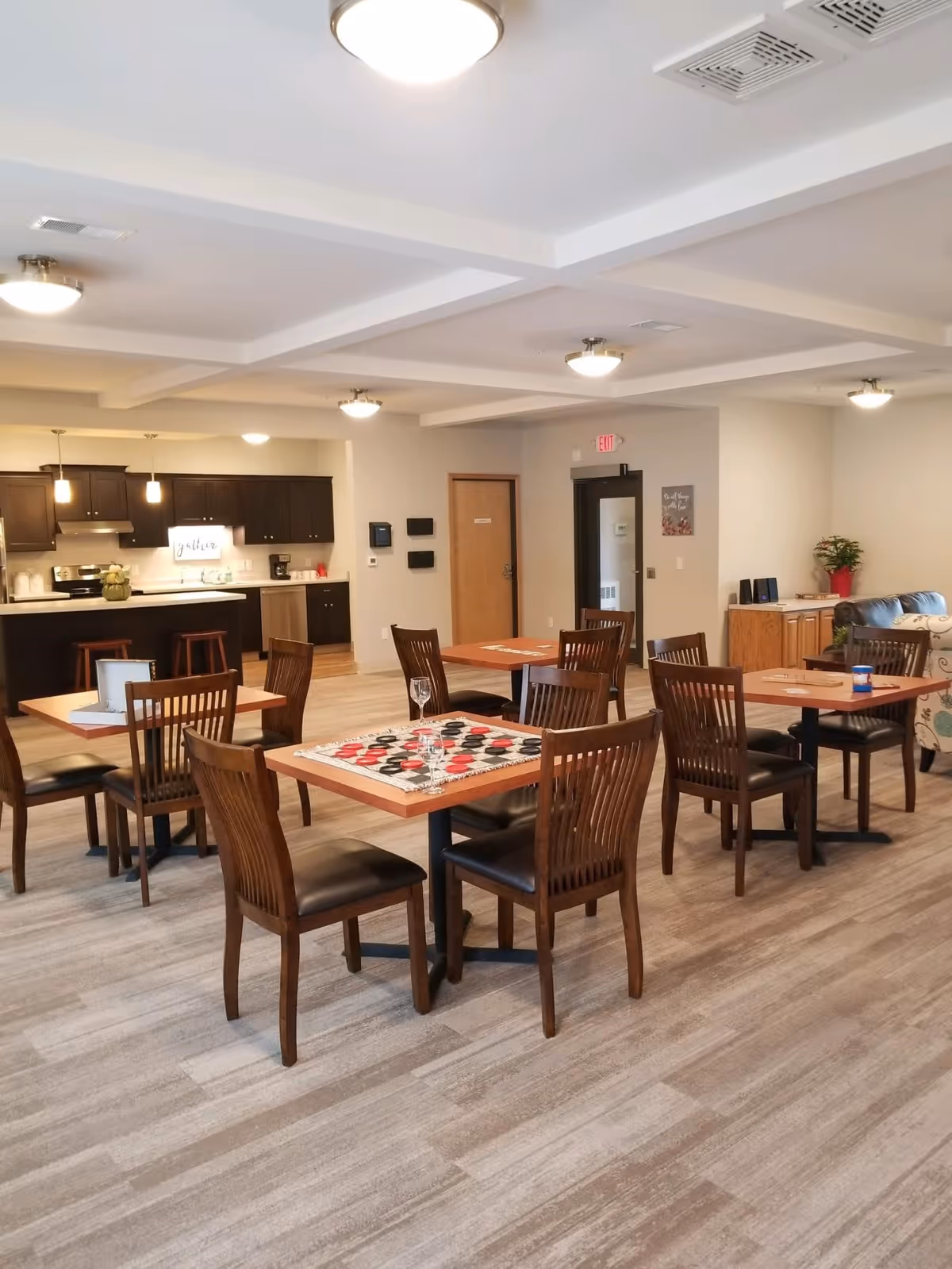 A spacious and well-lit common area with several wooden tables and chairs arranged for seating. One table has a checkers game set up with red and black pieces. In the background, there is a kitchen area with dark cabinets, a stove, a coffee maker, and a sign that says 'gather'. The room has light-colored walls and flooring, ceiling lights, and a door with an exit sign above it.