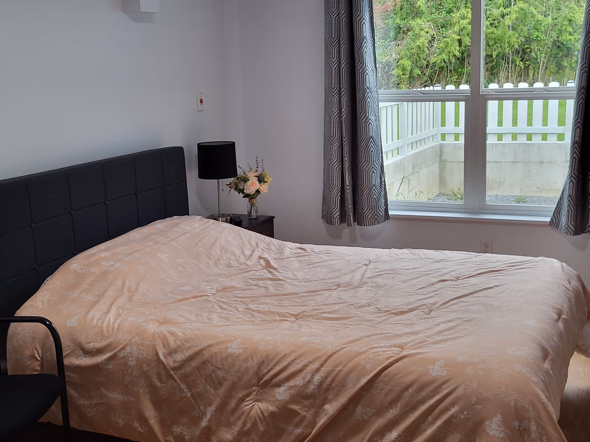 A bedroom with a large bed covered in a light peach floral-patterned comforter. There is a black upholstered headboard, a black chair partially visible on the left, a nightstand with a black lamp and a vase of flowers, and a window with patterned curtains showing a view of a white fence and greenery outside.