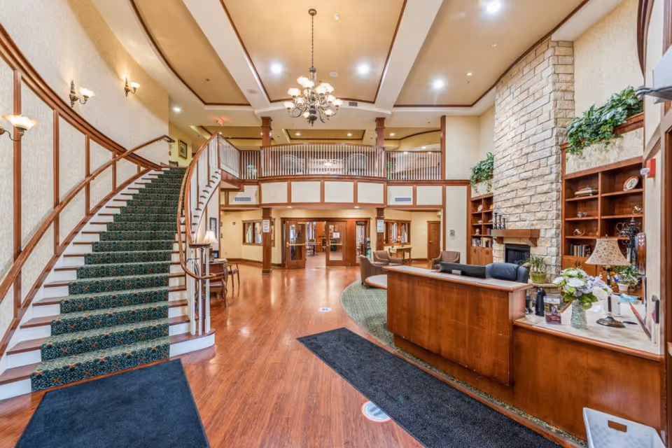 Spacious senior living facility lobby with a curved staircase on the left, wooden reception desk on the right, stone fireplace with built-in shelves, and chandelier lighting. The area features wood flooring, green carpet accents, and plants on shelves.
