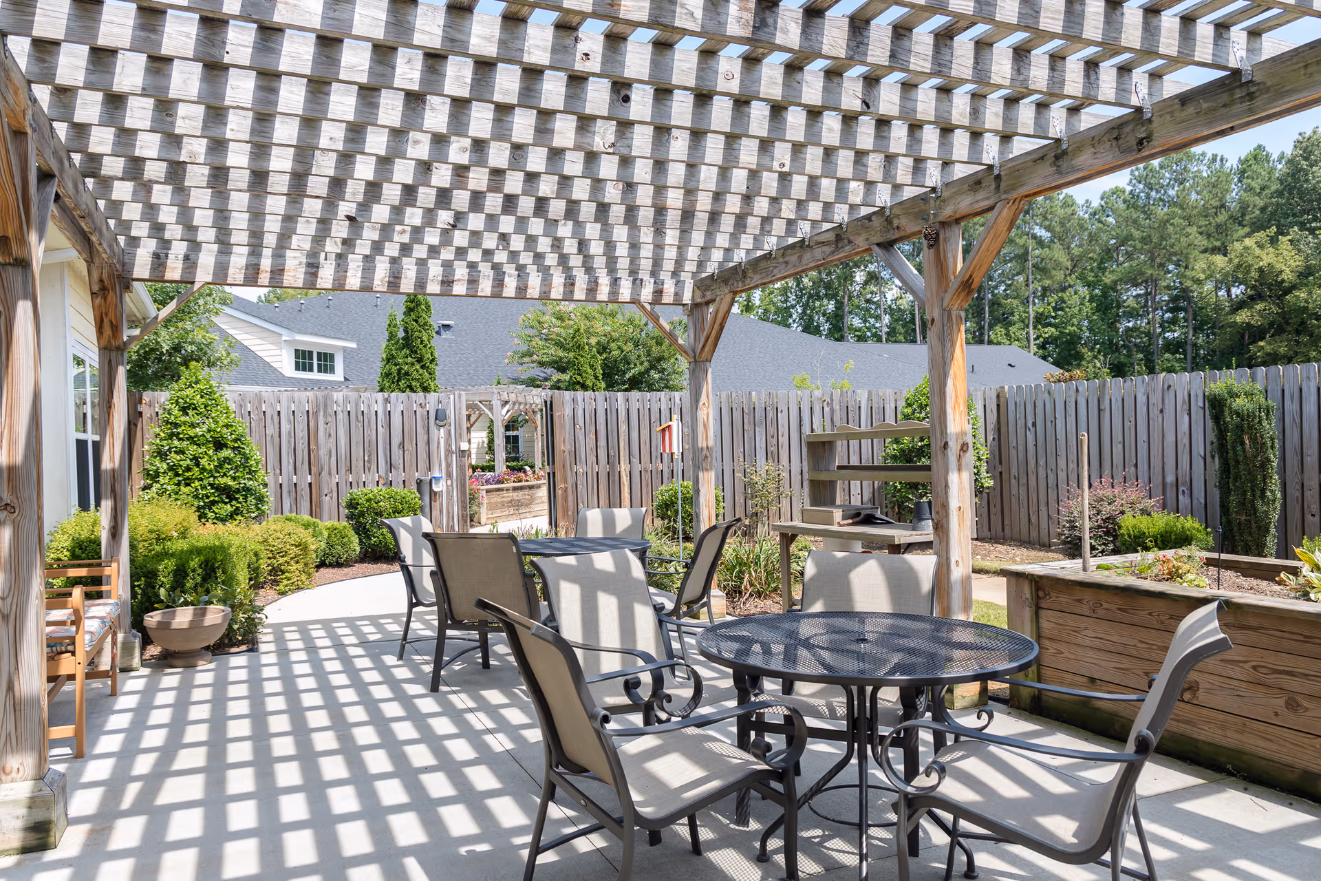 Outdoor patio area with a wooden pergola casting a checkered shadow on the concrete floor. There are several metal chairs with beige cushions around a round metal table. The patio is surrounded by a wooden fence and landscaped with green bushes and trees. A wooden bench and raised garden beds are also visible.