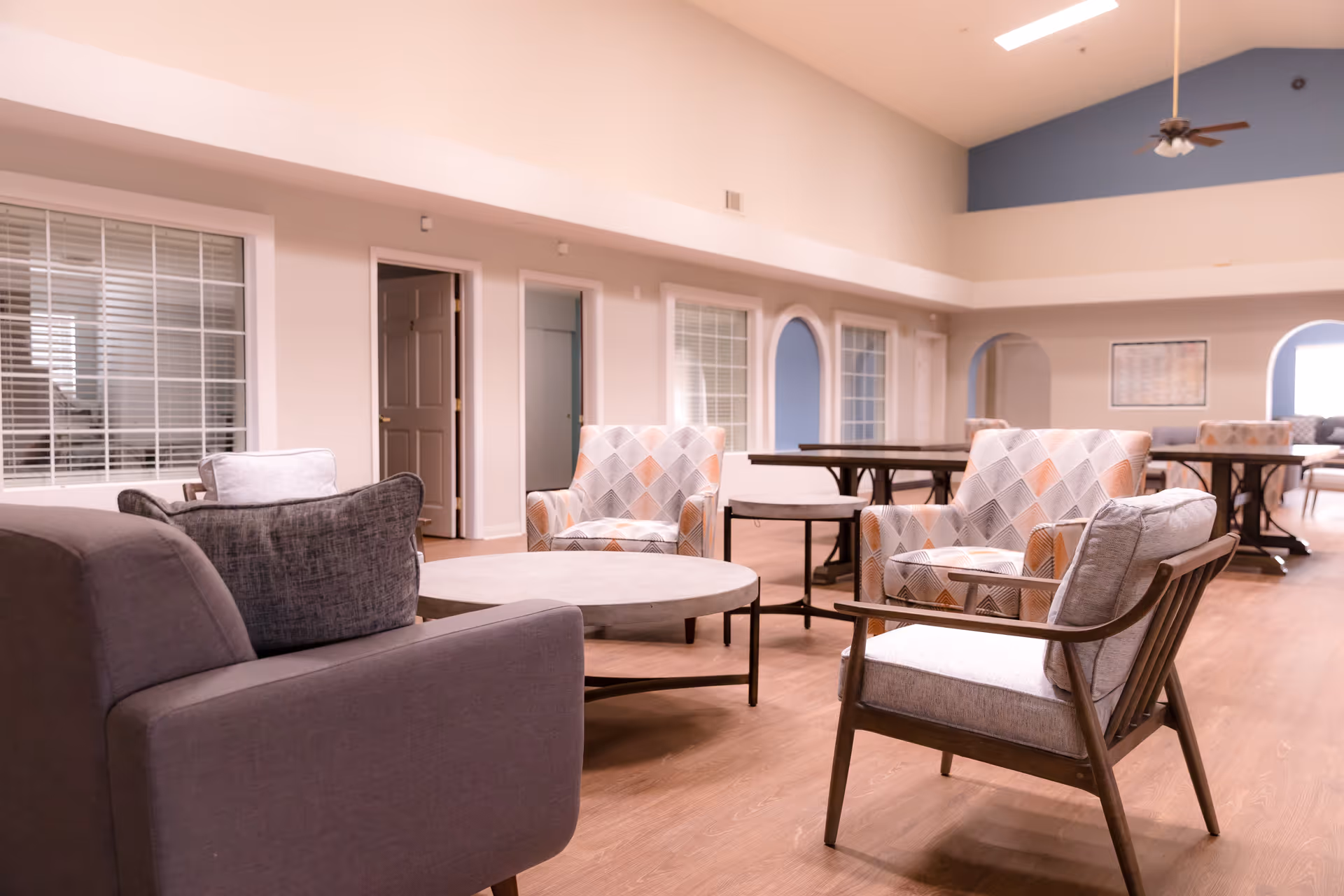 A spacious senior living common area with comfortable armchairs arranged around a low circular coffee table. The room features large windows with white blinds, light-colored walls, and a high ceiling with a ceiling fan. In the background, there are additional tables and chairs, and arched doorways leading to other rooms.