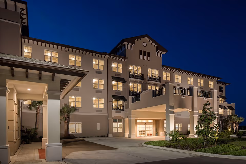 Well-lit multi-story senior living building front entrance at dusk with a porte-cochere and balconies.