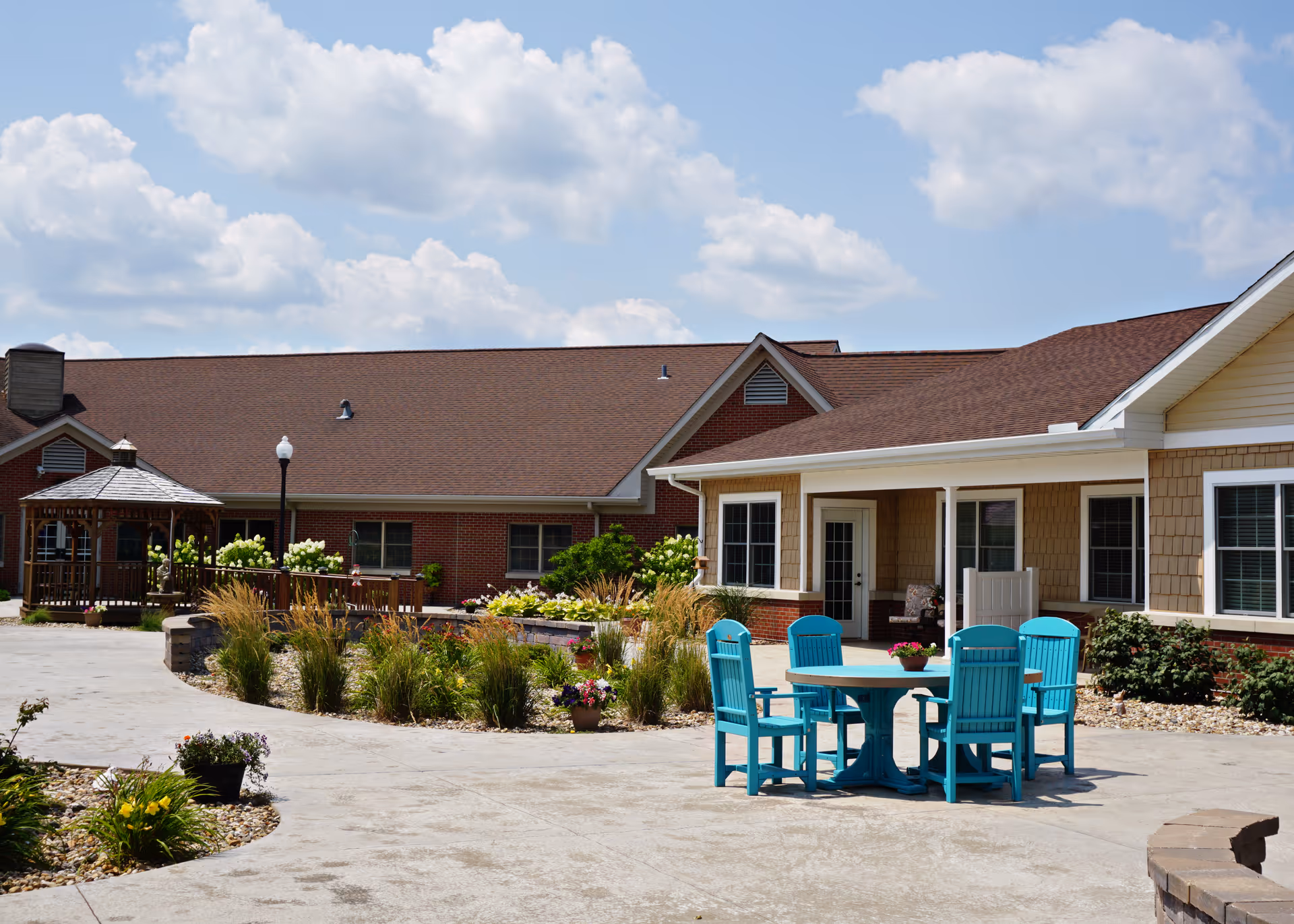 Outdoor patio area at Snyder Village Assisted Living featuring a round table with four bright blue chairs, surrounded by landscaped garden beds with various plants and flowers. The building has beige siding and brick accents with multiple windows and a covered porch area. A wooden gazebo and a lamppost are visible in the background under a partly cloudy sky.