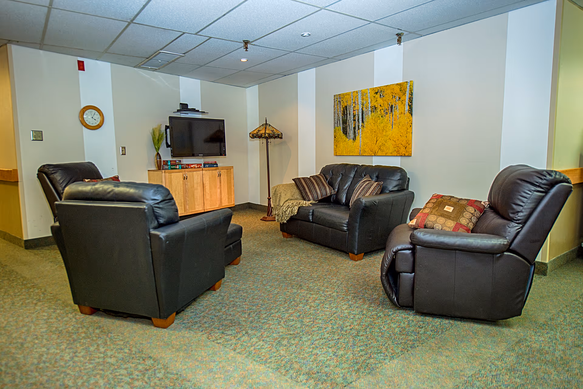 A cozy living room area with three black leather chairs and a matching loveseat arranged around a wooden cabinet with a flat-screen TV mounted above it. The room has a patterned carpet, a floor lamp, a wall clock, and a colorful painting of yellow trees on the wall.