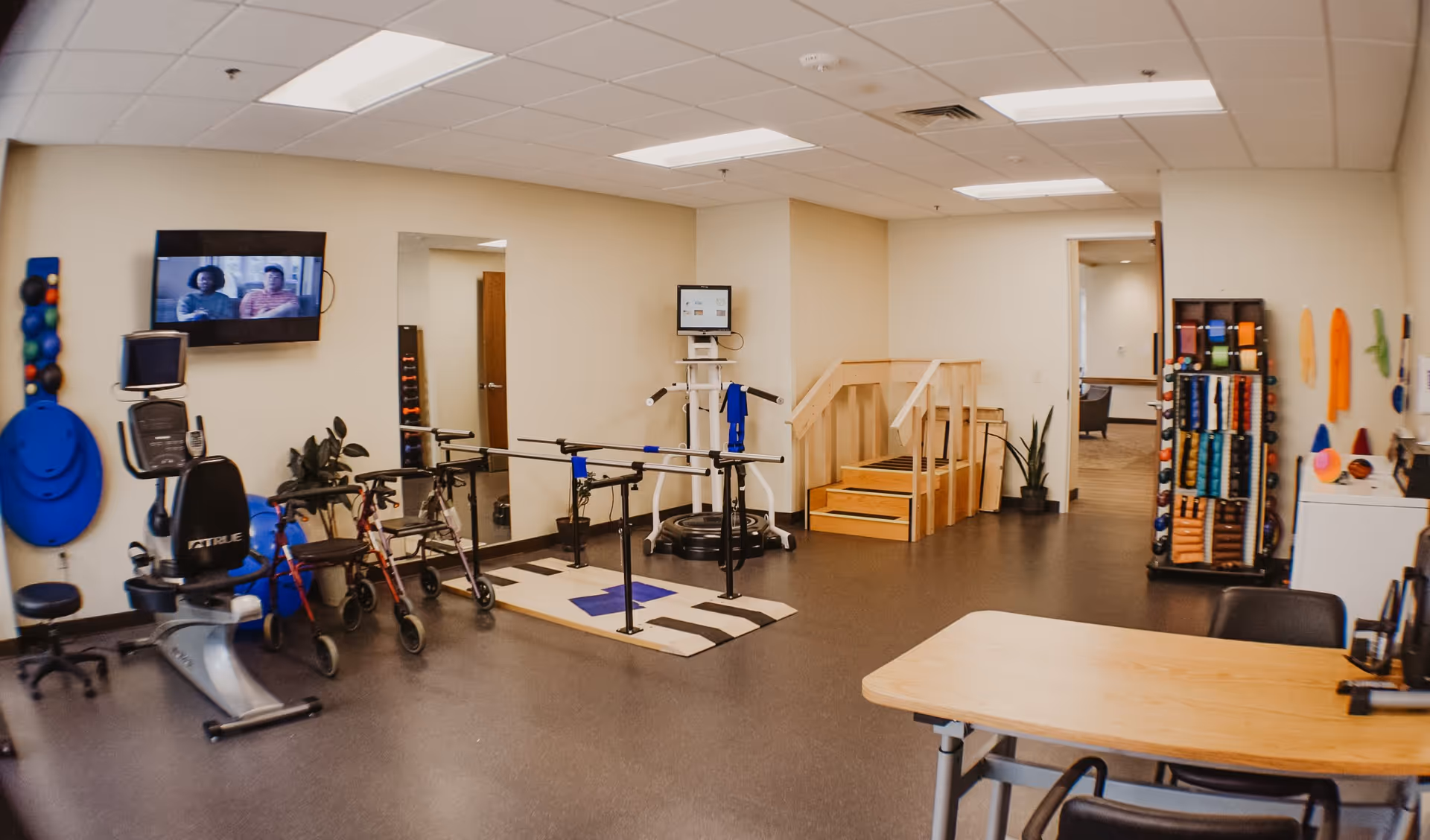 A physical therapy or rehabilitation room with exercise equipment including a stationary bike, parallel bars for walking practice, a set of stairs with handrails, and various therapy tools and weights organized on racks. A television is mounted on the wall, and there is a table with chairs in the foreground. The room has beige walls and a gray floor.