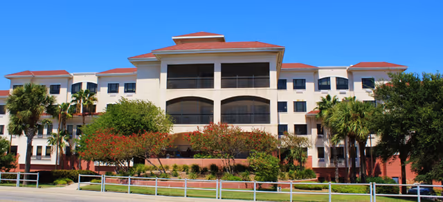 Exterior view of a multi-story senior living facility building with beige walls and red roofs, surrounded by palm trees and greenery under a clear blue sky.