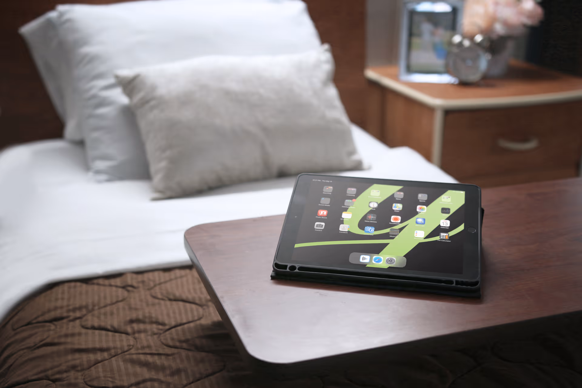 Tablet resting on a wooden overbed table in front of a made bed with pillows and a nightstand.