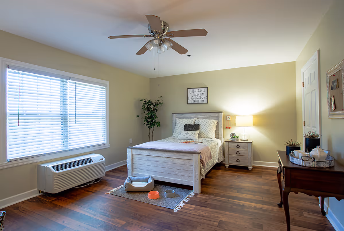 Bright furnished bedroom with a wooden bed, nightstand, ceiling fan and a large window with blinds.