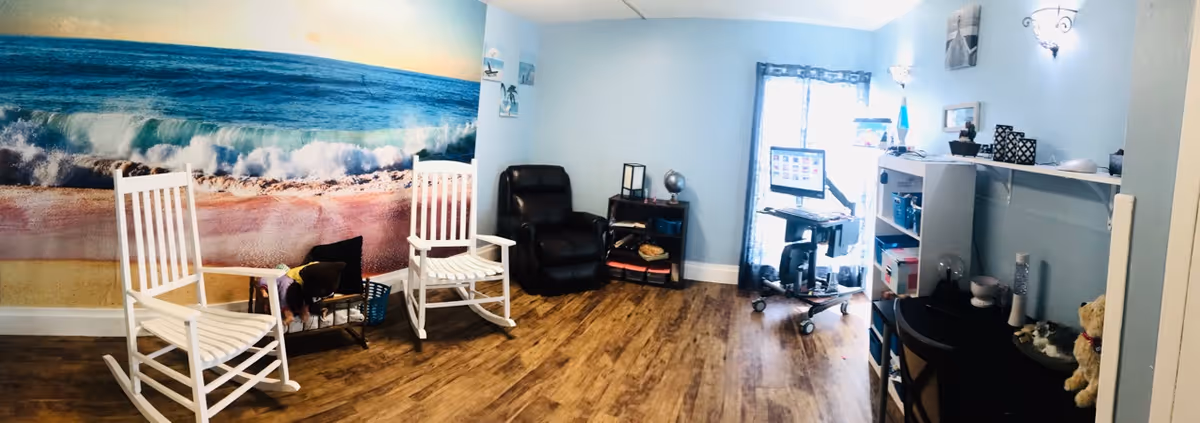 A cozy room with light blue walls and wood flooring featuring two white rocking chairs, a black recliner, a small shelving unit with books and decor, a computer workstation on wheels near a window with sheer curtains, and a desk with various items including a teddy bear. One wall has a large mural of an ocean wave crashing onto a sandy beach.