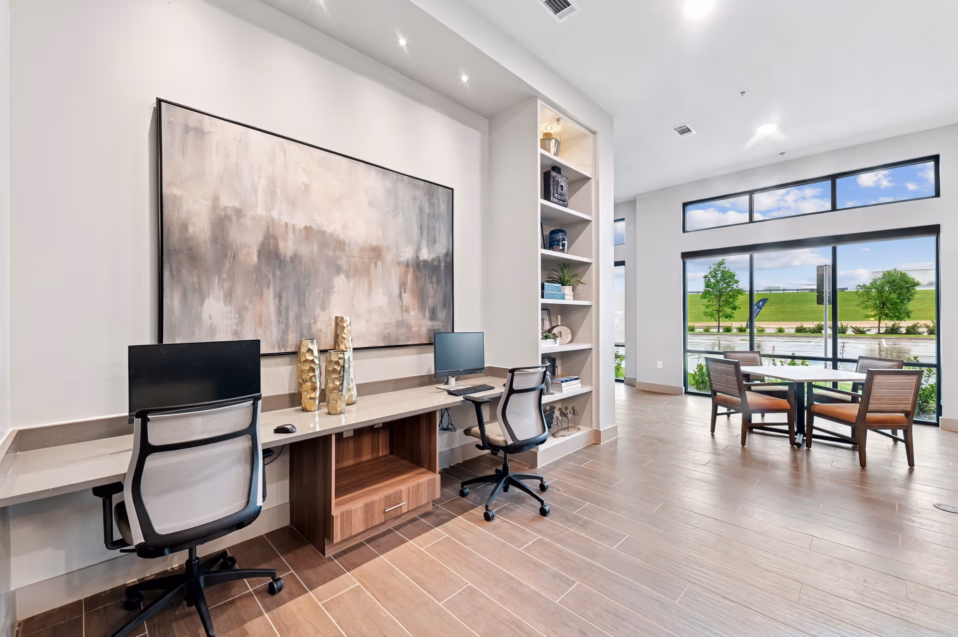 A modern interior space featuring a long desk with two office chairs and computer monitors. Above the desk is a large abstract painting. To the right, there is a built-in shelving unit with decorative items. Further in the background, there is a seating area with a table and four chairs near large windows that show a view of greenery and a blue sky outside.
