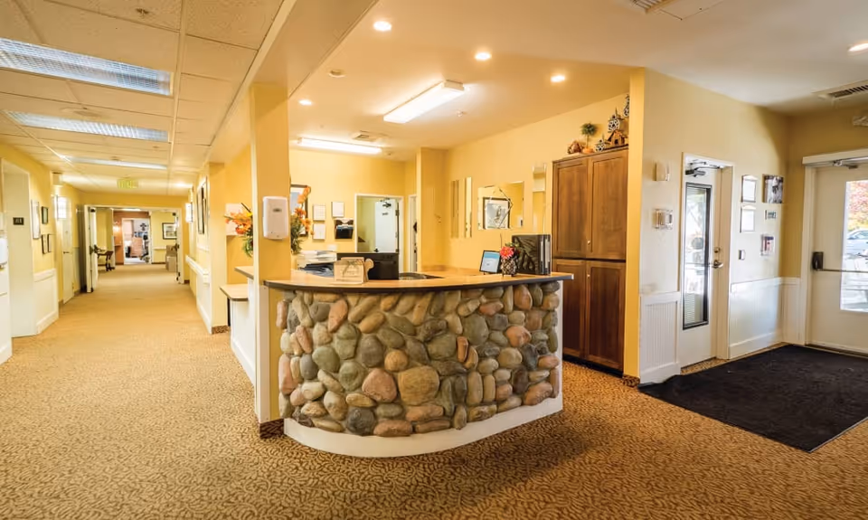 Reception area inside Lakeland Senior Living facility with a stone-faced curved desk, computer, and decorative items. The area is well-lit with yellow walls, carpeted floor, and a hallway extending to the left with doors and framed pictures on the walls. There are two glass doors on the right side leading outside.