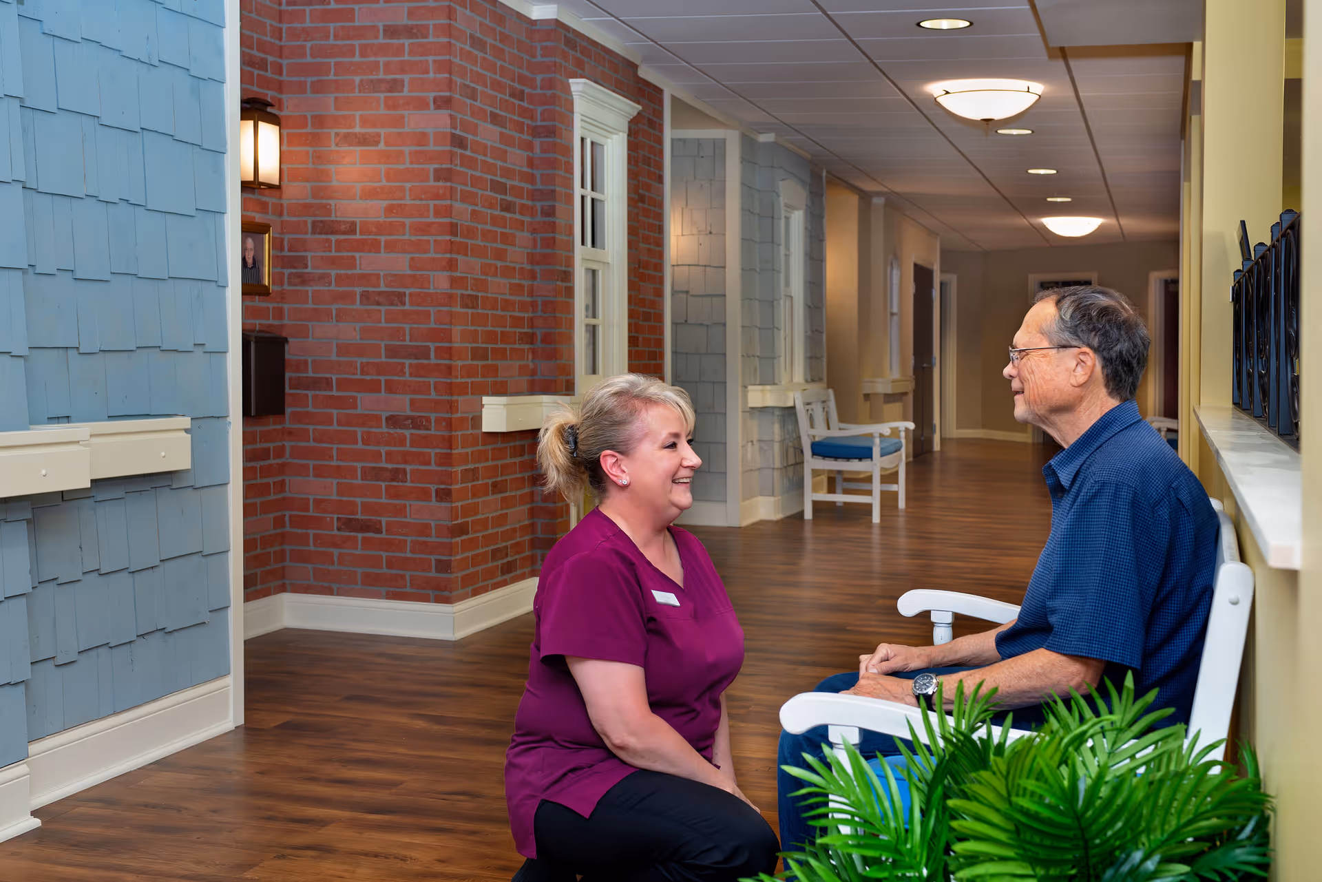 A woman in a purple uniform kneels and smiles while talking to an elderly man seated on a white bench in a hallway with wooden floors, brick and shingle walls, and ceiling lights.