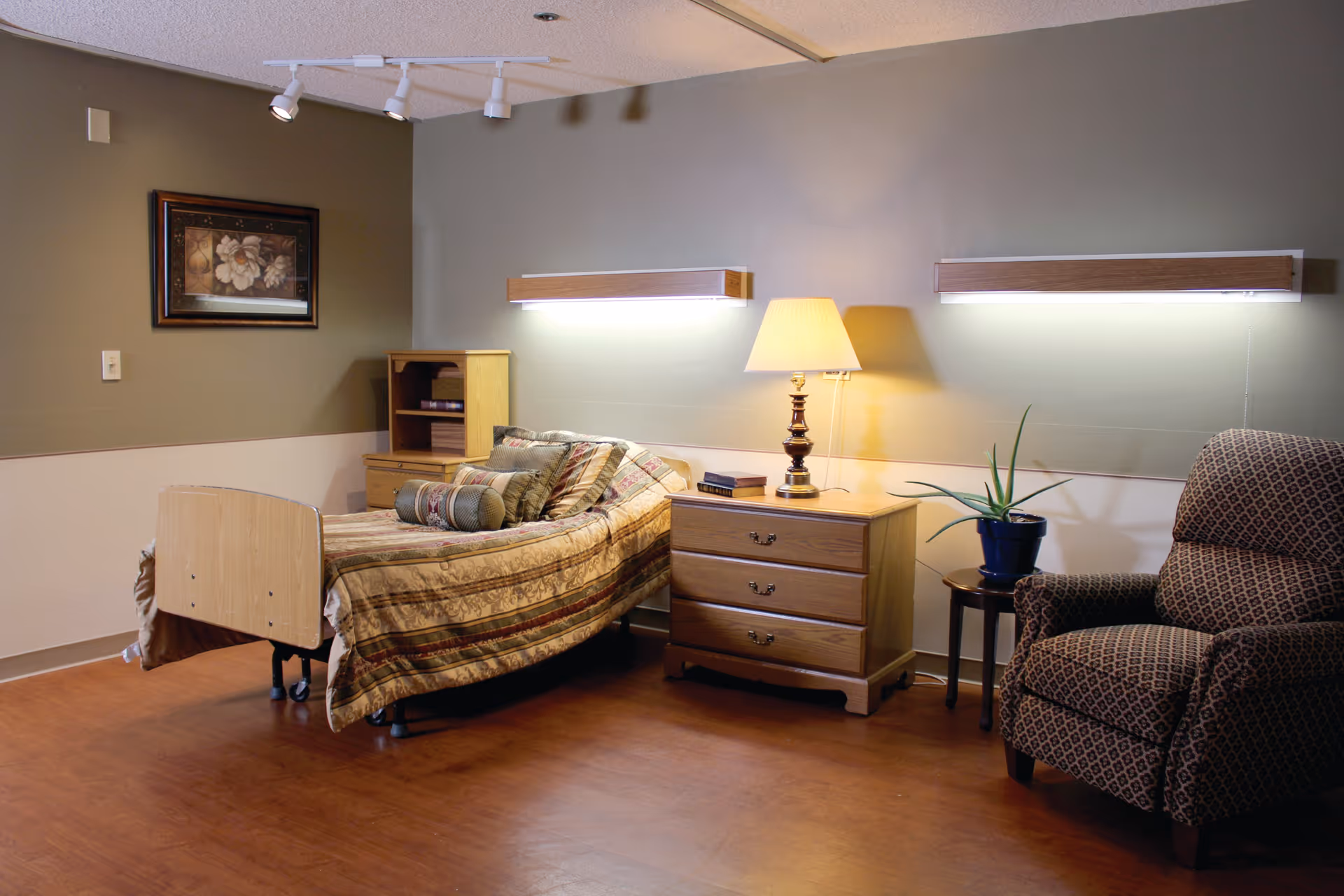 A cozy room with a single bed covered in a patterned bedspread and multiple pillows. Next to the bed is a wooden nightstand with a lamp and some books. There is a small wooden shelving unit beside the bed. A patterned armchair and a small round table with a potted plant are also visible. The walls are painted in two tones, with a framed floral picture hanging on one side. The floor is wooden, and two wall-mounted lights illuminate the room.