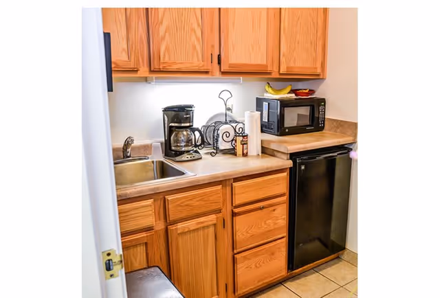 Small kitchen area with wooden cabinets, a stainless steel sink, a coffee maker, a paper towel holder, a microwave, a small black refrigerator, and a bowl with bananas on the countertop.