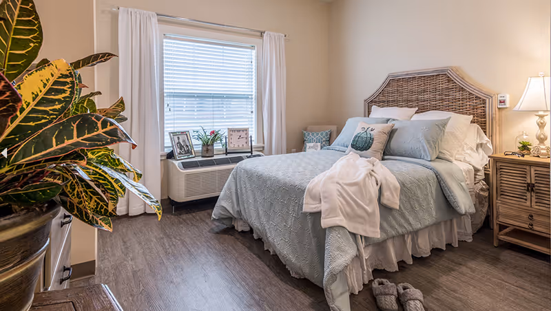 Sunny bedroom with a wicker headboard bed dressed in light blue linens, a bedside table and lamp, slippers on the floor, and a potted plant by the window.