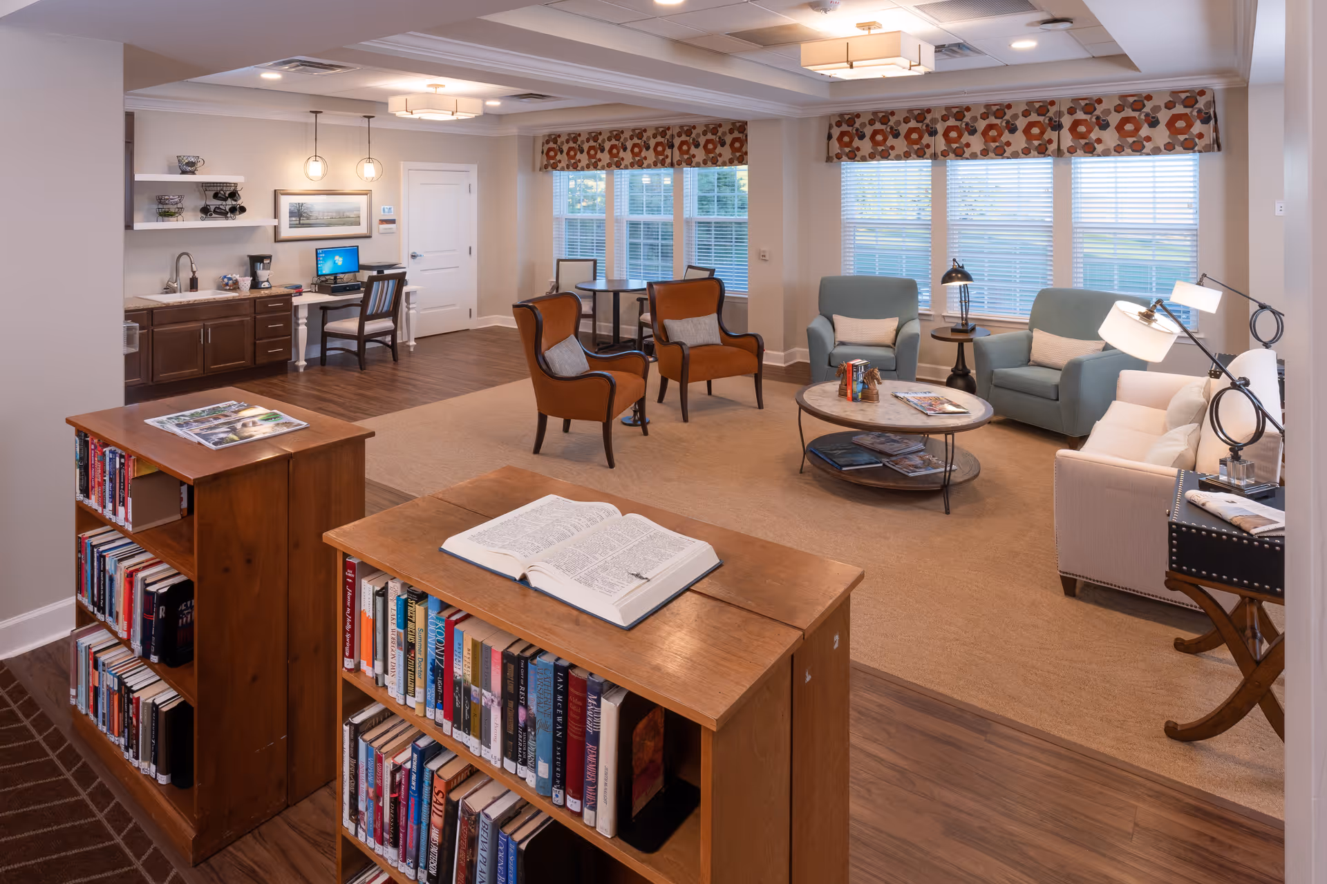 A cozy living room area in The Culpeper facility featuring a carpeted seating space with two blue armchairs, a beige sofa, two brown chairs, and a round coffee table. The room has large windows with patterned valances, wooden bookshelves filled with books, a small desk with a computer, and warm lighting fixtures on the ceiling and lamps.
