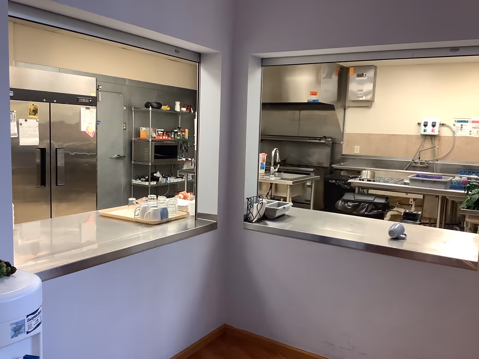 Commercial stainless-steel kitchen seen through two pass-through serving windows with counters, appliances, and shelving.