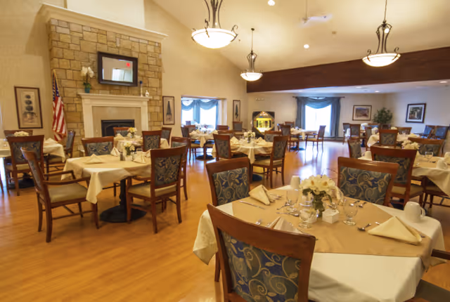 A spacious dining room in Oak Creek Place Assisted Living Community with multiple tables covered in beige tablecloths and set with napkins, glasses, and floral centerpieces. The room features wooden chairs with patterned cushions, a stone fireplace with a mirror above it, and large windows with blue curtains allowing natural light to enter. Elegant hanging light fixtures illuminate the room.