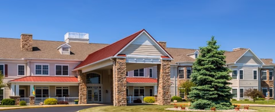 Exterior view of a senior living facility named Waterford Crossing with a large covered entrance supported by stone pillars, beige siding, red metal roofing accents, multiple windows, and a well-maintained lawn with a large evergreen tree in front.