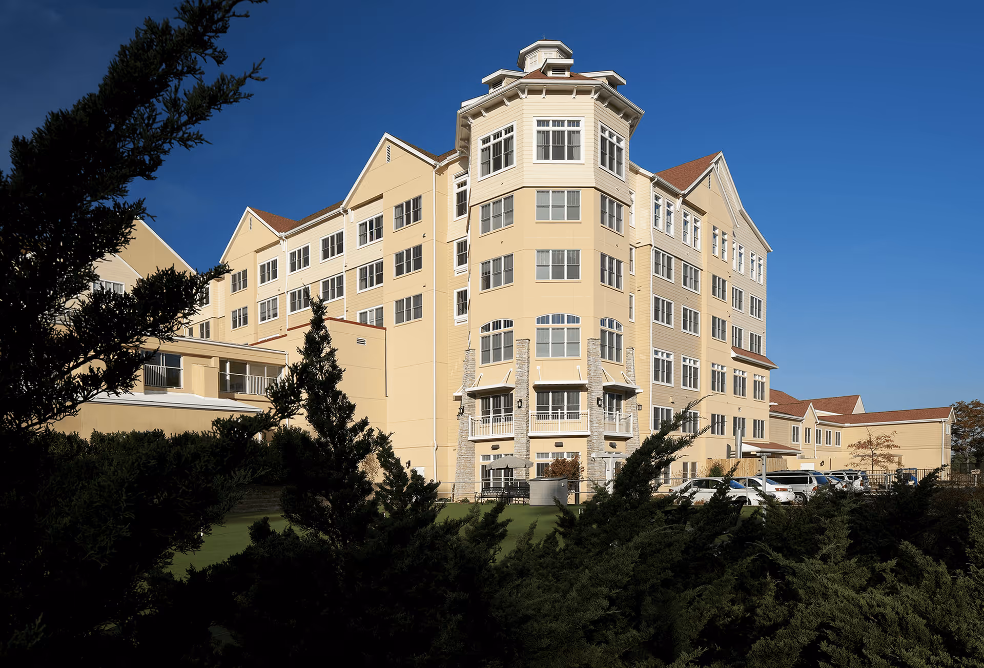 Exterior view of a large multi-story beige retirement community building with many windows, surrounded by green bushes and trees under a clear blue sky.