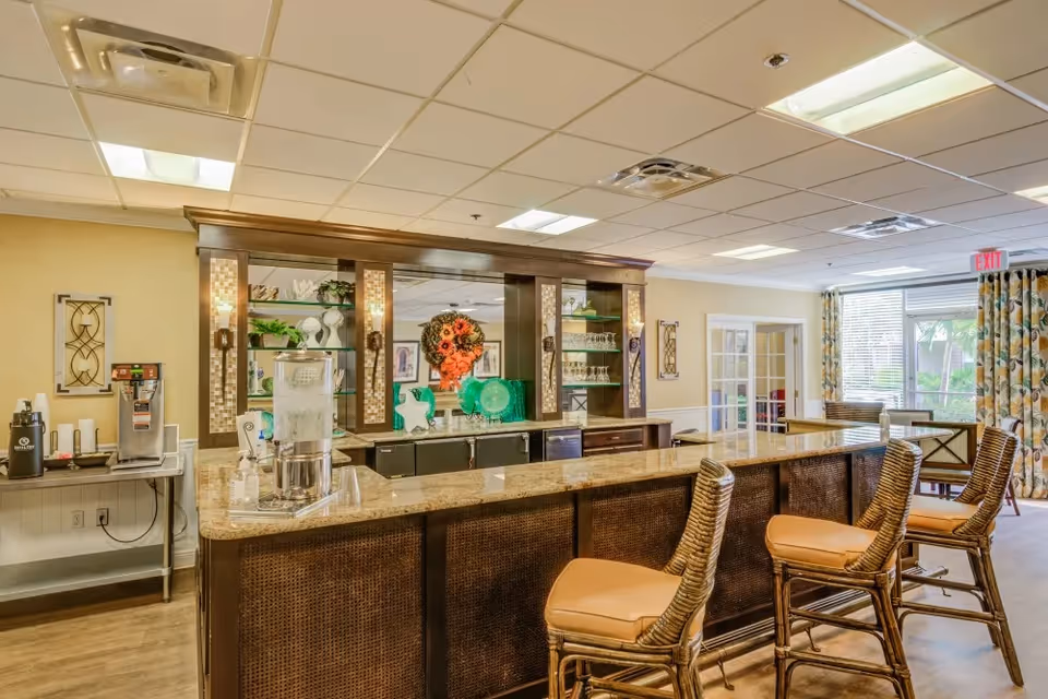 Interior view of a senior living facility's dining or common area featuring a long granite countertop bar with four wicker bar stools with tan cushions. Behind the bar is a wooden cabinet with glass shelves displaying decorative items and glassware. To the left, there is a beverage station with a coffee dispenser and water container. Large windows with patterned curtains allow natural light into the room.