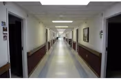 Long institutional hallway in a senior living facility with doors on both sides, handrails, and fluorescent ceiling lights.