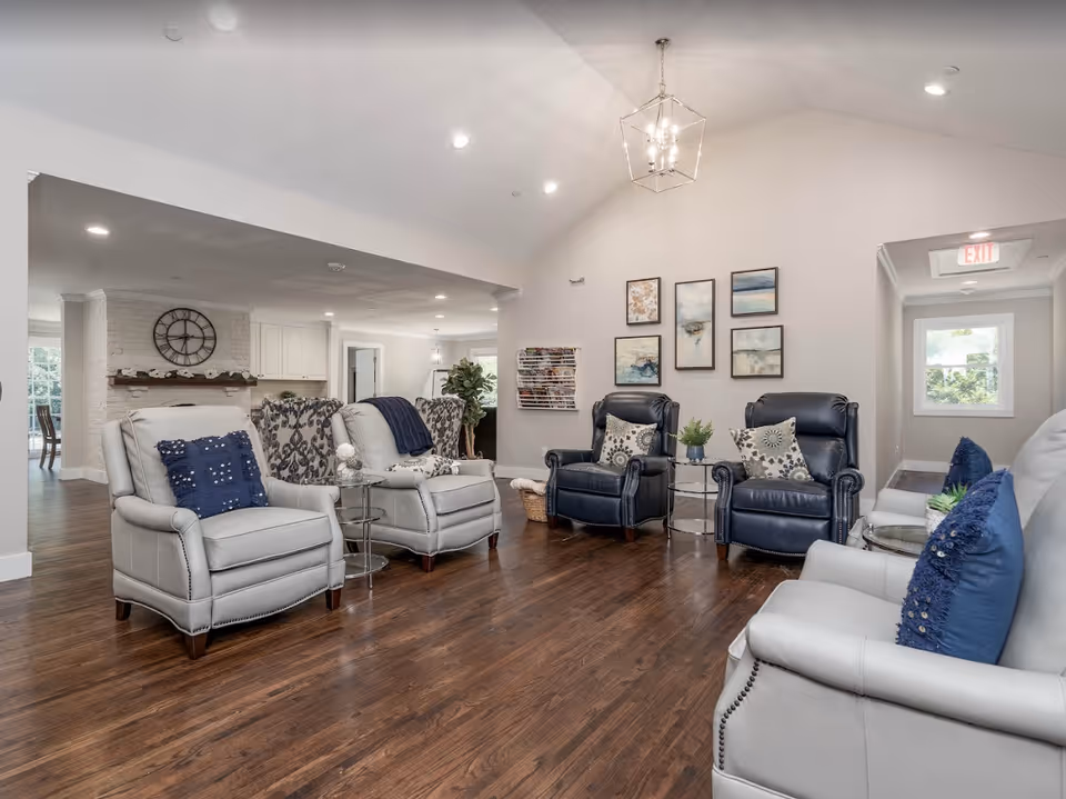 A bright and spacious living room area with several comfortable armchairs arranged in a semi-circle on a wooden floor. The chairs are upholstered in light gray and dark blue leather, each adorned with decorative pillows. The walls are painted light beige and decorated with a series of framed abstract paintings. A modern chandelier hangs from the vaulted ceiling, and a magazine rack is mounted on one wall. In the background, a white brick fireplace with a large clock above it is visible, along with a glimpse of a dining area and a hallway with an exit sign.