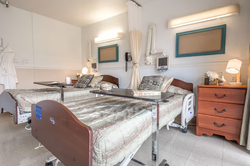 Two hospital-style patient beds in a shared room with curtains, furniture, and medical equipment.