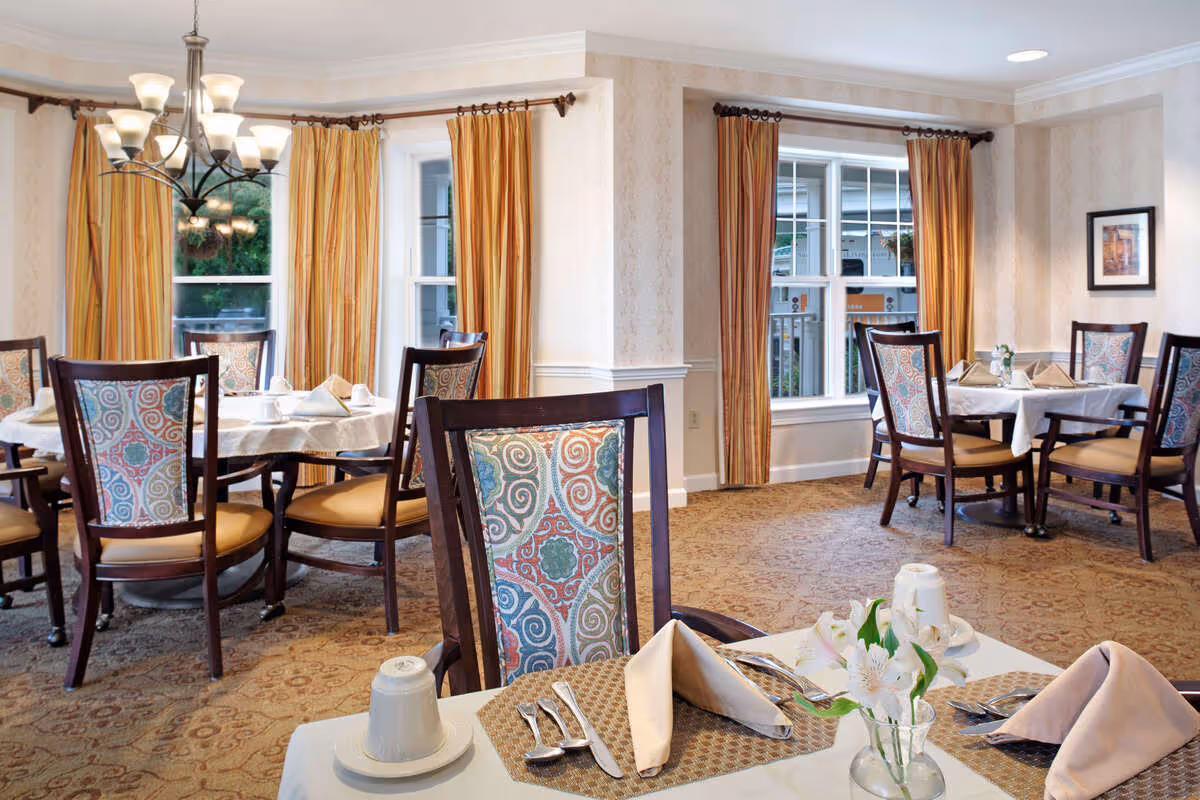 Bright dining room with round tables, patterned upholstered chairs, place settings, and large windows with striped curtains.