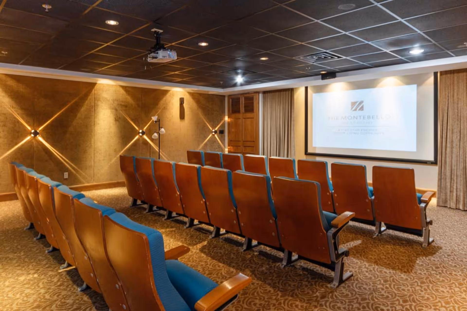 A small theater-style room with rows of wooden chairs with blue cushions facing a large projection screen. The walls are decorated with warm lighting fixtures, and the ceiling has a black grid pattern with recessed lights and a ceiling-mounted projector.