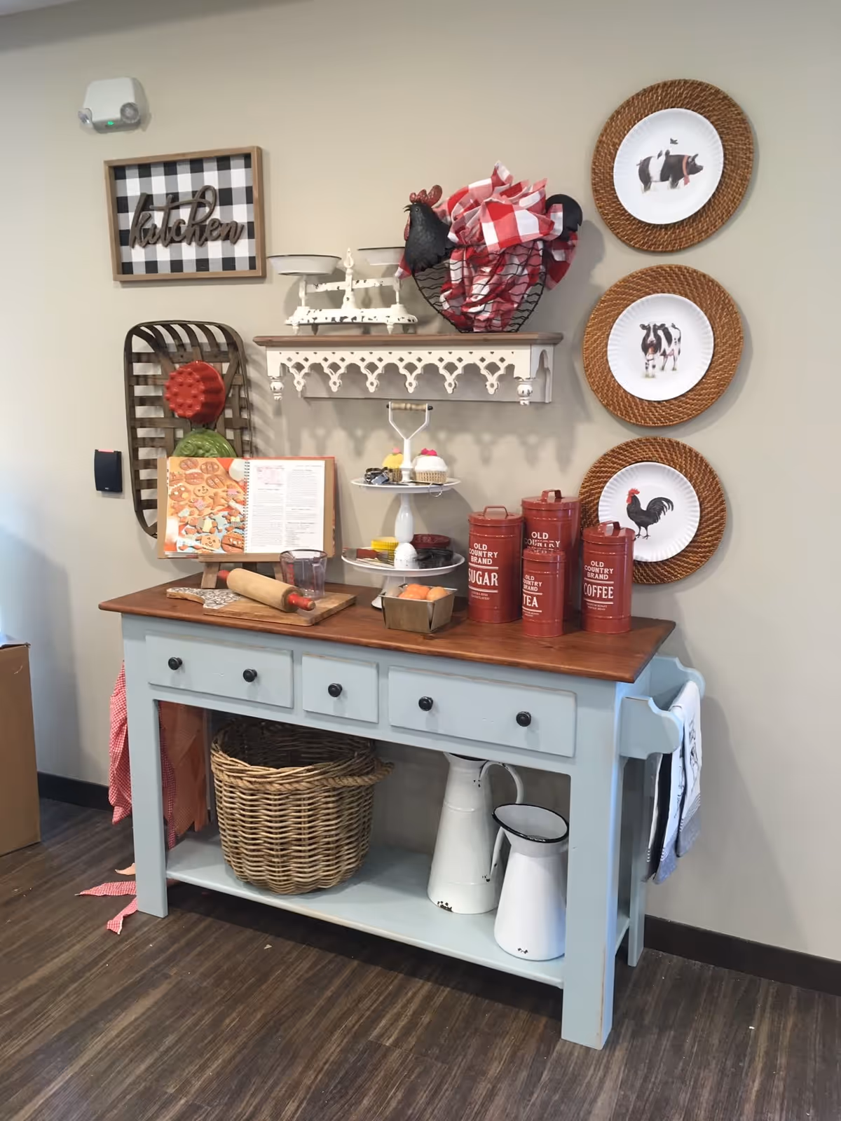 A rustic kitchen corner with a light blue wooden table featuring three drawers and a wooden top. On the table are red canisters labeled sugar, tea, and coffee, a tiered stand with cupcakes, a rolling pin, a cookbook on a stand, and a small basket with eggs. Above the table is a decorative white shelf holding a vintage scale and a wire basket with red and white checkered cloth. Three decorative plates with farm animal images are mounted on the wall to the right, and a framed sign reading 'kitchen' hangs on the wall to the left. Below the table is a large wicker basket and two white enamel pitchers. The floor is dark wood, and a towel hangs on the side of the table.
