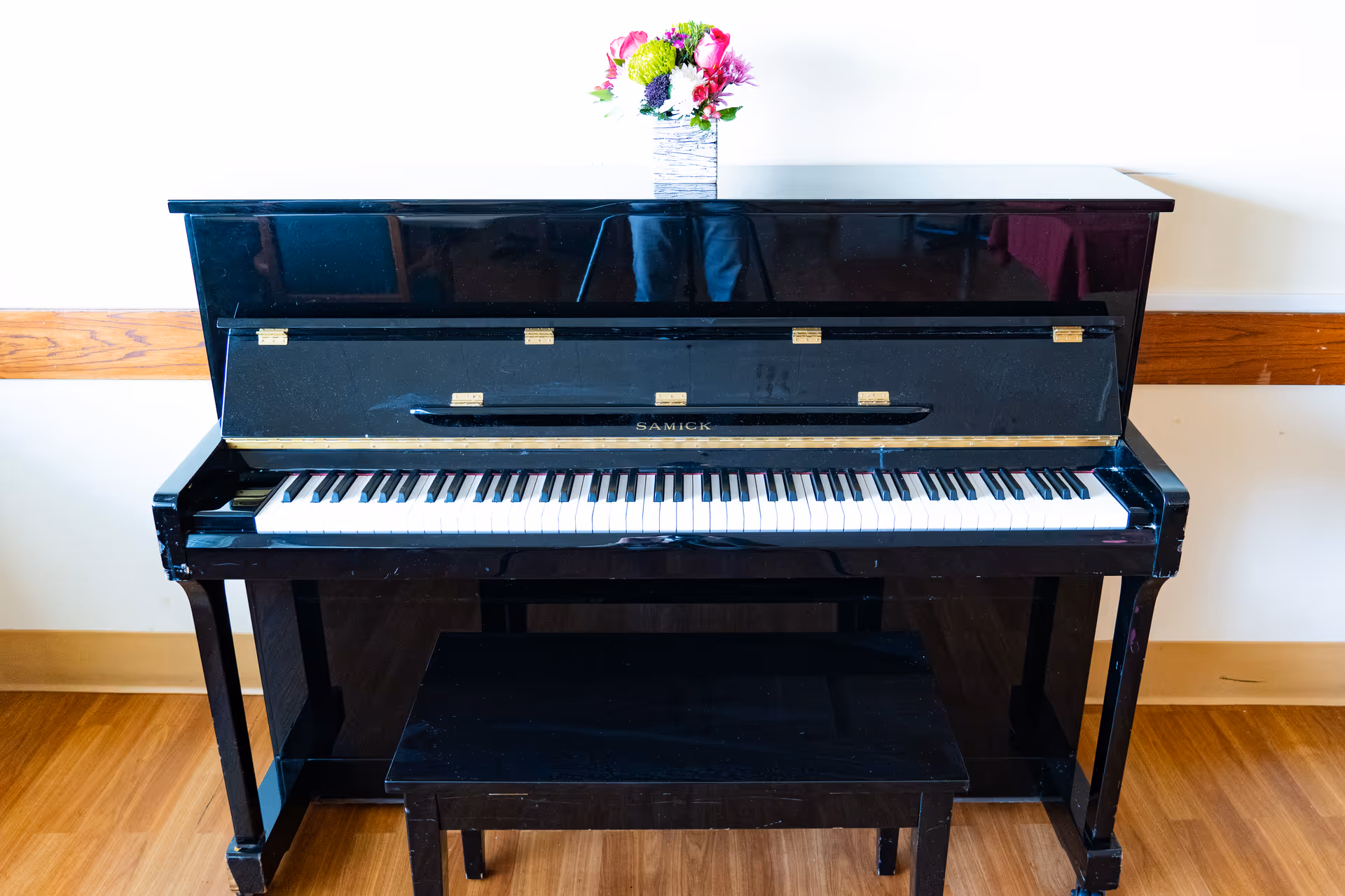 A black upright piano with a matching black bench in front of it. On top of the piano is a small vase with colorful flowers. The piano is placed against a white wall with a wooden chair rail and a wooden floor beneath it.