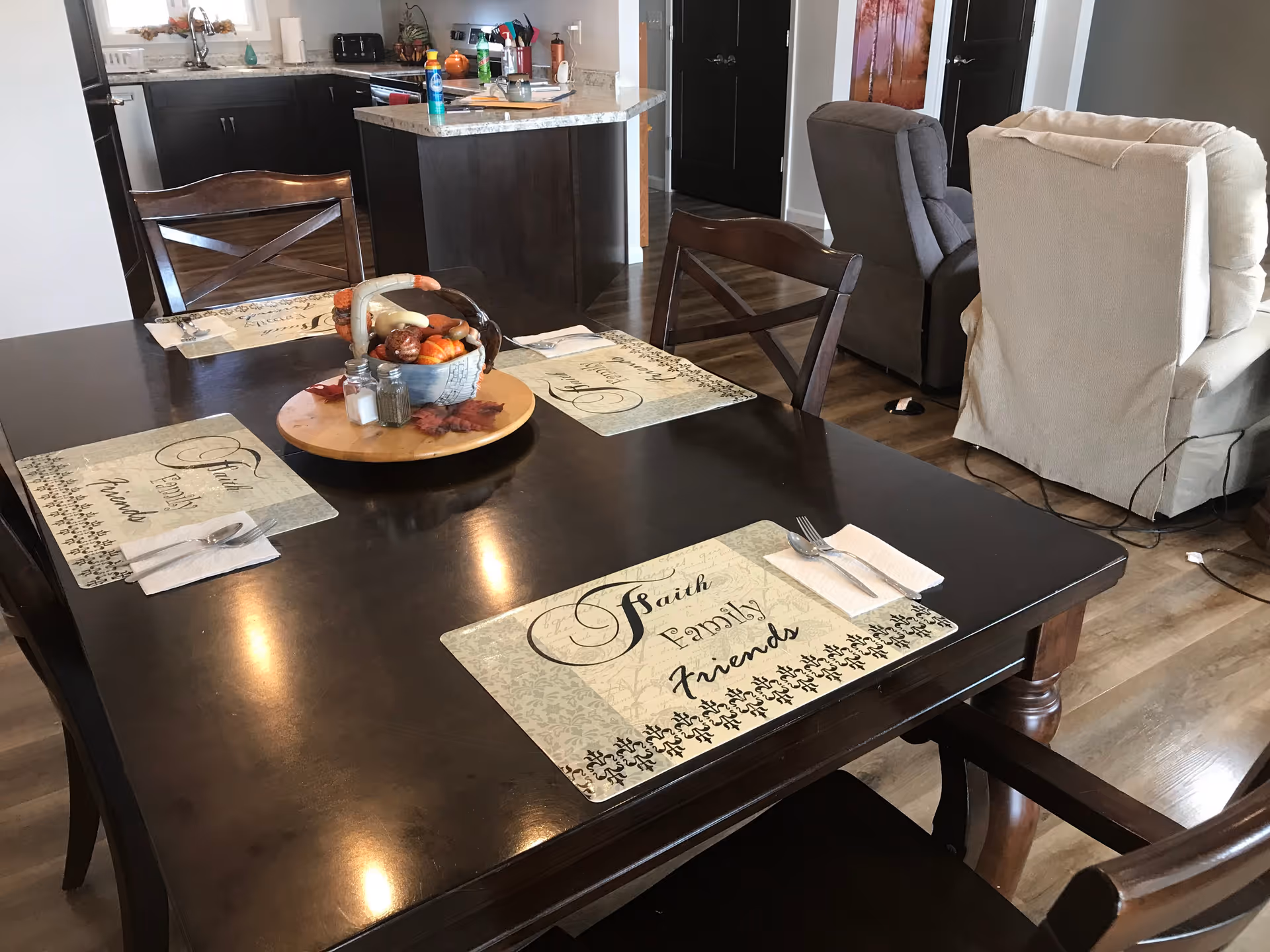 Dark wooden dining table with placemats and a fruit centerpiece in an open-plan kitchen and living area with recliners in the background.