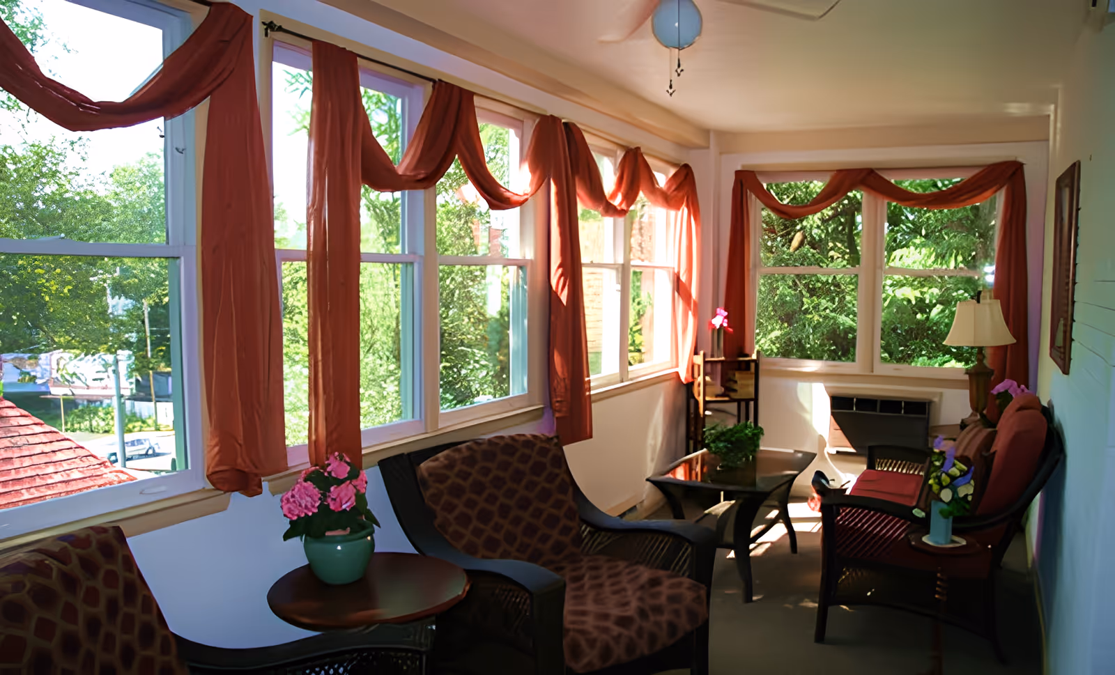A bright sunroom with large windows draped with rust-colored curtains. The room is furnished with cushioned wicker chairs, small tables holding potted plants and flowers, and a table lamp. Outside the windows, green trees and a red roof are visible.