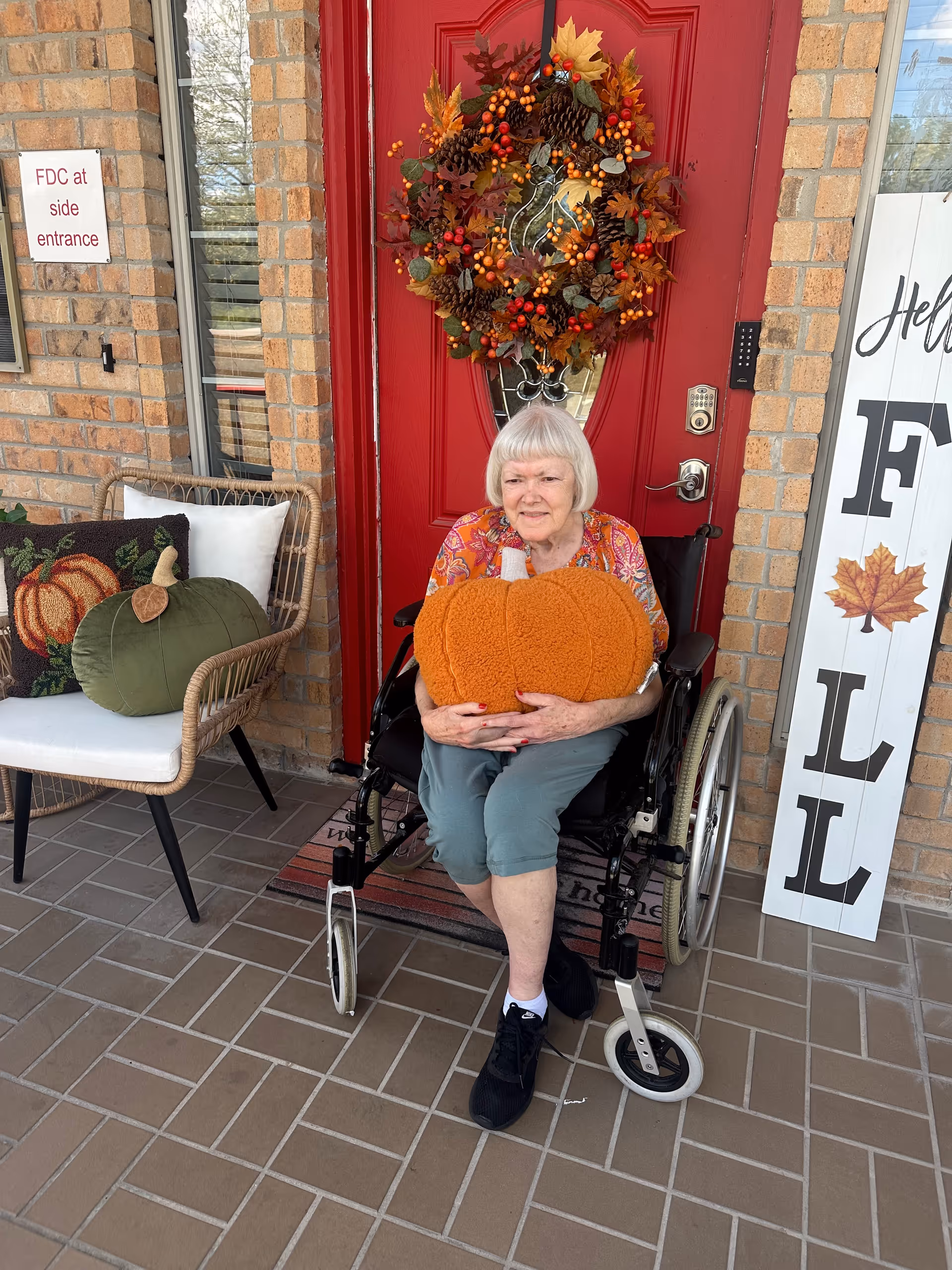 An elderly woman sitting in a wheelchair on a tiled porch in front of a red door decorated with a fall-themed wreath. She is holding a large orange pumpkin-shaped cushion. To her left is a wicker chair with fall-themed pillows, and to her right is a white wooden sign with the word 'FALL' and a maple leaf decoration. A sign on the brick wall reads 'FDC at side entrance.'