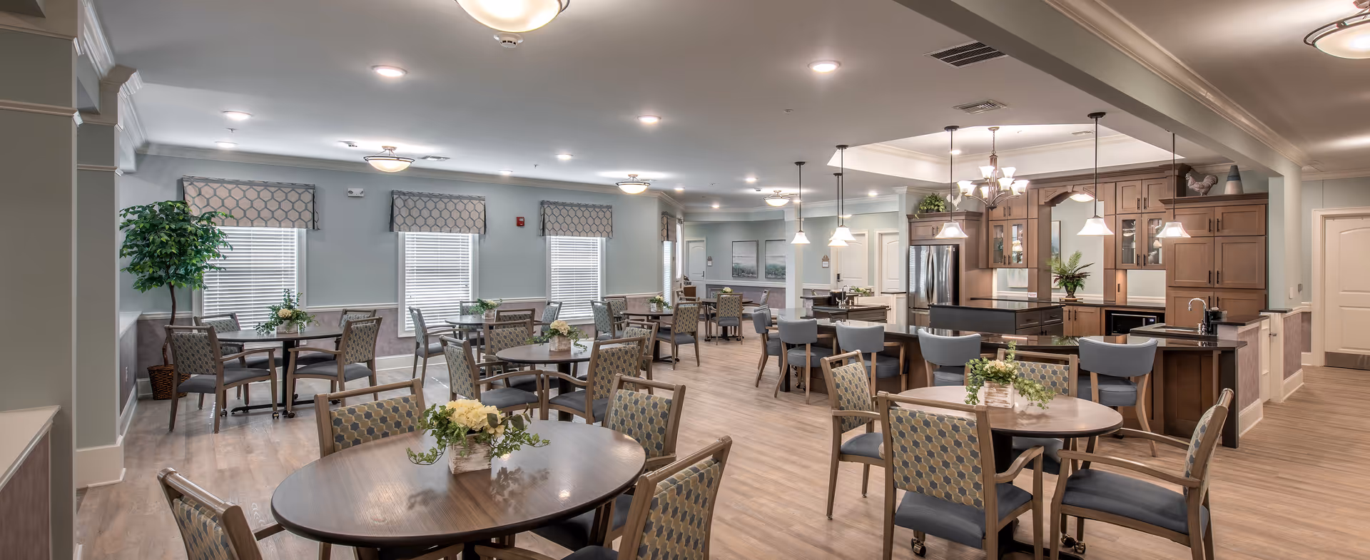 A spacious and well-lit dining area in a senior living facility featuring multiple round tables with patterned chairs, floral centerpieces, large windows with patterned valances, and a kitchen area with wooden cabinetry, a refrigerator, and pendant lighting.