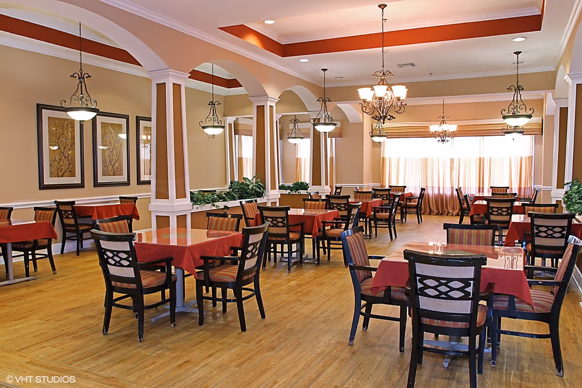 A spacious dining room with multiple tables covered in red tablecloths and surrounded by wooden chairs with patterned cushions. The room features warm beige walls, decorative arches with columns, framed artwork, hanging light fixtures, and large windows with sheer curtains allowing natural light to fill the space. The floor is a light wood finish.