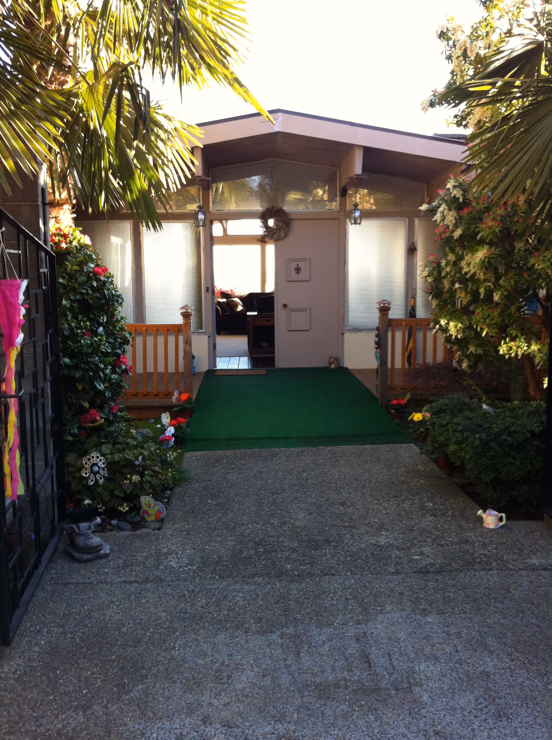 Entrance to a building with a green carpeted ramp leading to a door. The entrance is flanked by plants and flowers on both sides, with palm leaves hanging overhead. The door has a wreath and windows on either side with blinds. Inside, a couch and table are visible through the door.