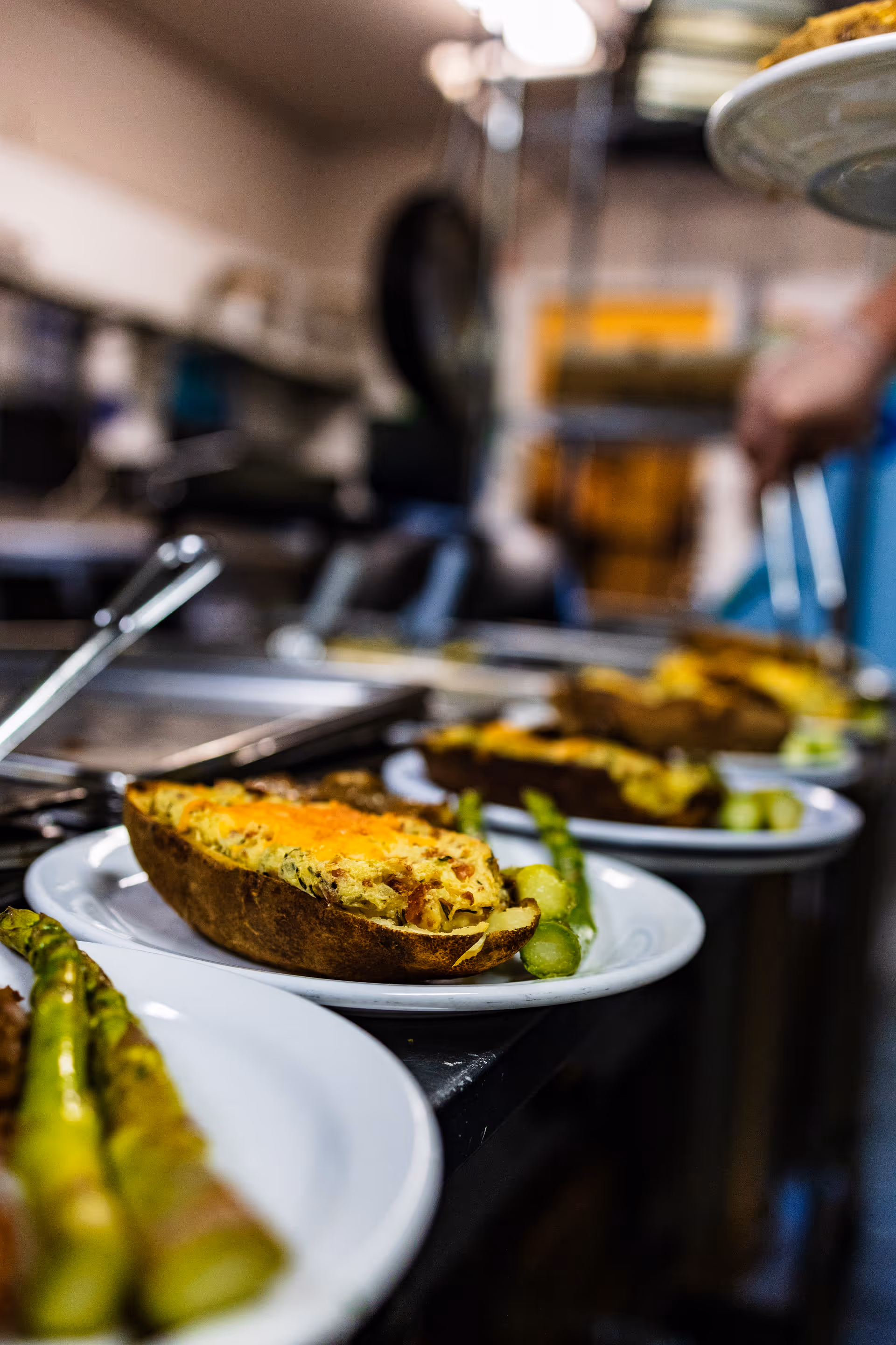 Close-up view of plates with stuffed baked potatoes and asparagus spears lined up on a kitchen counter, with a blurred background of a person serving food in a commercial kitchen.