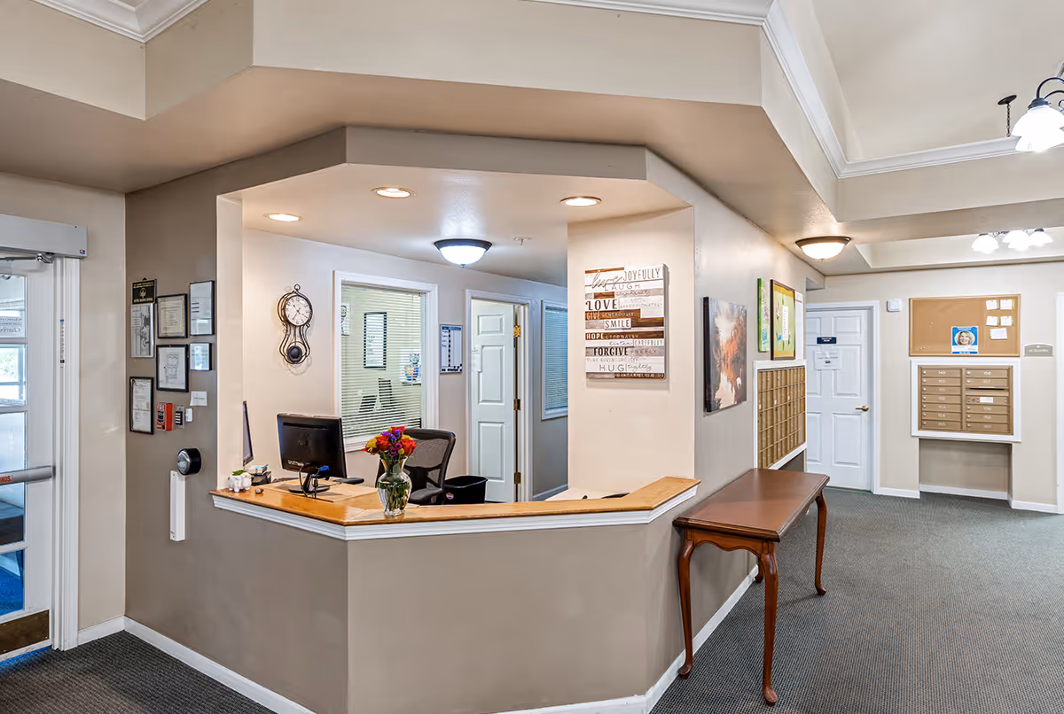 Reception area of Rolling Meadows Senior Living with a wooden counter, a computer, a vase with flowers, wall clock, framed certificates, and a bulletin board with mailboxes in the background.